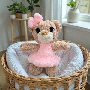 May include: A handmade crochet teddy bear with a pink dress and bow. The bear is light brown with black eyes and a white muzzle. It sits in a wicker basket lined with a white, fluffy blanket. A charming, soft toy.