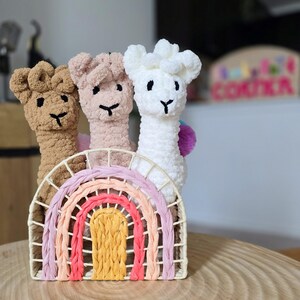 May include: Three crocheted llama toys in brown, pink, and white, standing in a rainbow-colored woven basket.