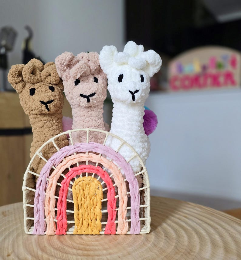 May include: Three crocheted llama toys in brown, pink, and white, standing in a rainbow-colored woven basket.