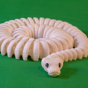 May include: A coiled, off-white snake sculpture with a detailed, segmented body and a small, detailed head. The snake is positioned on a solid green background, creating a striking contrast.