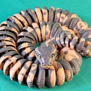 May include: A coiled, articulated snake toy with a detailed design. The toy features segments in shades of tan and gray, with a snake head at the center. The snake has green eyes and is posed on a green surface.