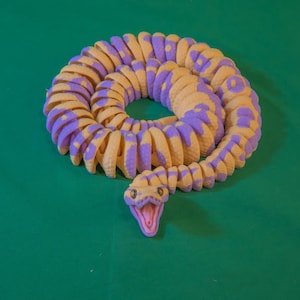 May include: A coiled, articulated snake toy in shades of yellow and purple, with an open mouth revealing a pink interior. The toy is positioned on a solid green surface.