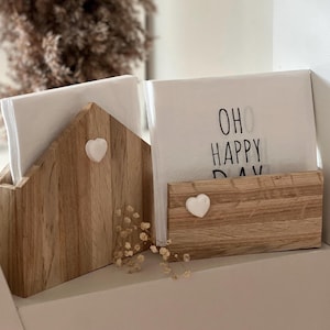 Op de afbeelding: Houten servetthouder met twee witte servetten. De servetthouder is in de vorm van een huis met een wit hart aan de voorkant. Een servet heeft de tekst "Oh Happy Day" erop gedrukt.
