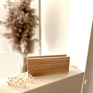 Könnte beinhalten: Ein rechteckiger Schreibtisch-Organizer aus Holz mit hellem Holzmaserungsfinish. Der Organizer hat zwei Fächer und eine dünne weiße Akzentlinie entlang der Oberkante. Im Hintergrund befindet sich ein Trockenblumenarrangement.