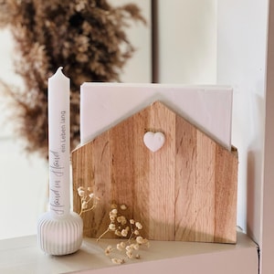 Op de afbeelding: Een houten servetthouder in de vorm van een huis met een wit hart aan de voorkant. Een witte kaars met de tekst "Hand in Hand ein Leben lang" staat op de voorgrond.