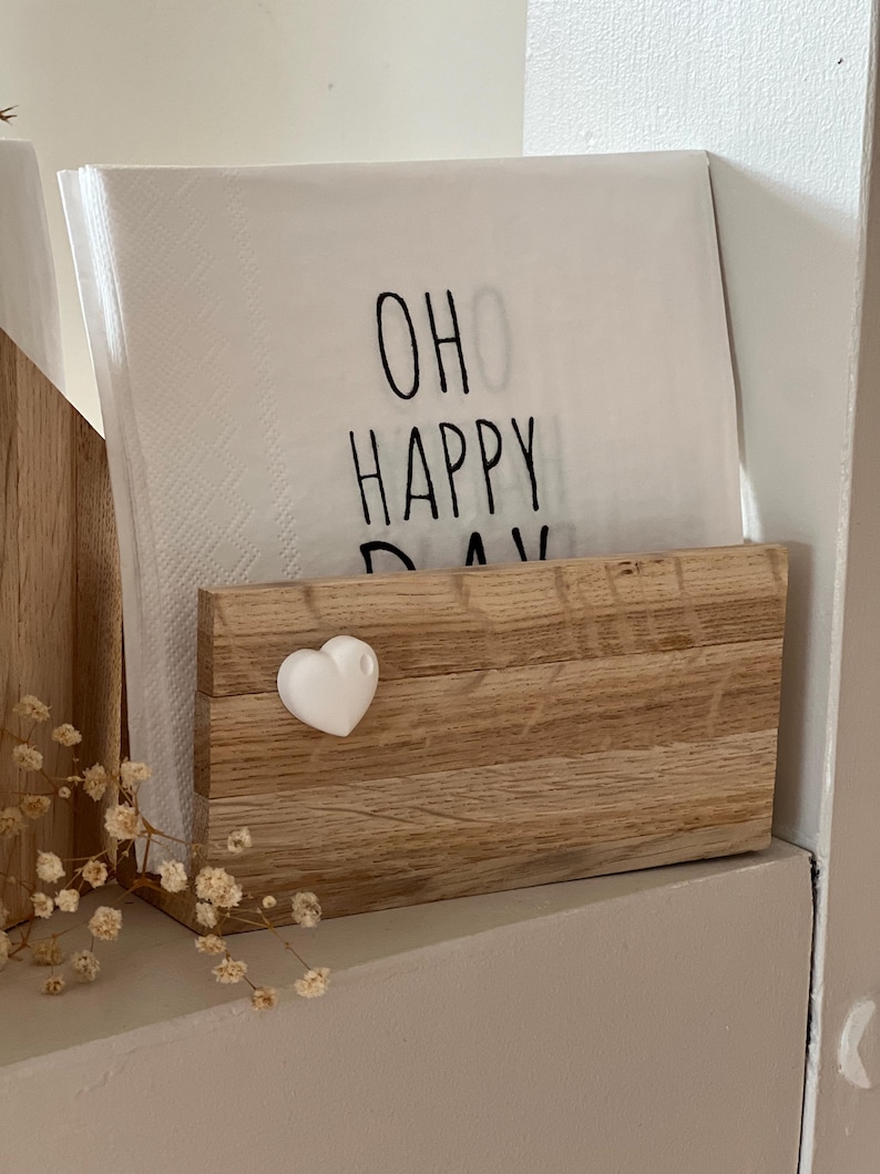 Op de afbeelding: Een houten servetthouder met een wit hart aan de voorkant. De servetthouder heeft een stapel witte servetten met de tekst "Oh Happy Day" erop gedrukt.
