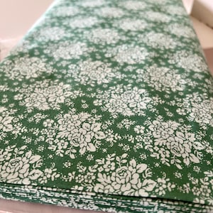 Könnte beinhalten: Ein Stapel grüner Stoffe mit einem weißen Blumenmuster. Der Stoff zeigt ein detailliertes Muster aus weißen Blumen und Blättern auf einem tiefgrünen Hintergrund. Der Stoff ist mit "Audrey Ocean Green" beschriftet.