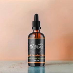 May include: A dark brown glass bottle with a black dropper top and a black label. The label has white text that reads "Old Fashioned Lounge" and "Agave Sunrise Bitter". The bottle contains 2 fl oz (60 ml) of liquid.