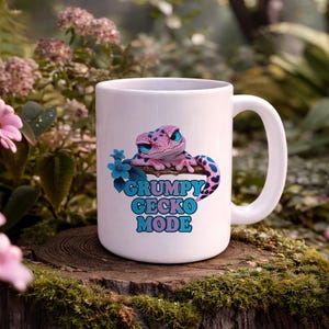 Könnte beinhalten: Weißer Keramikbecher mit einem Cartoon-Gecko-Design. Der Gecko ist rosa und blau, mit blauen Augen, und der Text lautet "GRUMPY GECKO MODE". Der Becher steht auf einer Holzoberfläche, umgeben von Grün.