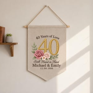 May include: A beige fabric wall banner featuring embroidered text that reads 40 Years of Love, Still Hand in Hand, Michael & Emily, and 12.08.1986. The design includes a large gold number 40 and a cluster of pink and white roses with green leaves.
