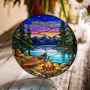 Könnte beinhalten: Ein runder Buntglas-Sonnenfänger mit einer Landschaft aus Kanu, Lagerfeuer, Kiefern, See und Bergen unter einem Sternenhimmel. Der Text "COUNTRY ROADS, TAKE ME HOME 2026" ist oben auf der Glasscheibe aufgedruckt.