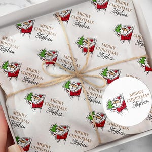 May include: A white gift box containing tissue paper printed with a repeating pattern of Santa Claus holding a Christmas tree. The tissue paper is tied with twine. The text "MERRY CHRISTMAS Stephen" is printed on the paper and a round sticker.