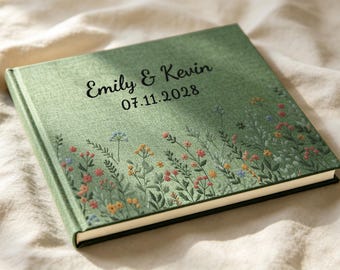 May include: A sage green fabric-covered photo album featuring a printed wildflower meadow design at the bottom. The cover displays the black text "Emily & Kevin" above the date "07.11.2028" in a dark, elegant script and sans-serif font combination.