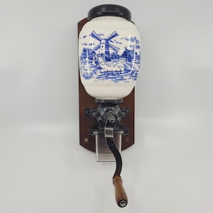 Op de afbeelding: Een vintage koffiemolen met een witte keramische container met een blauwe windmolenscène. De molen is gemonteerd op een houten plank en heeft een zwart metalen mechanisme met een houten handvat. Een klassiek keuken decoratie-item.