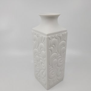 May include: White porcelain vase with an intricate embossed floral design. The vase has a square base and a slightly flared opening.