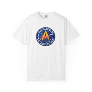 May include: A white short-sleeve t-shirt featuring a circular Starfleet Command United Federation of Planets emblem on the chest. The logo displays a yellow delta insignia with a red swoosh against a dark blue starry background with white stars.