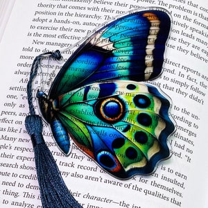 May include: A blue and green butterfly bookmark with a tassel. The butterfly has a detailed design with a mix of colors and patterns. The bookmark is made of clear acrylic and has a text overlay.