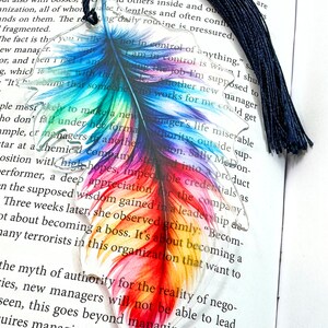 May include: A colorful feather bookmark with a rainbow design. The bookmark is made of clear acrylic and has a blue tassel. The bookmark is placed on top of an open book.