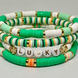 May include: A set of four bracelets with green, white, and orange beads. The bracelets are stacked on top of each other and spell out the word "LUCKY" with white beads and gold lettering. A gold shamrock charm is on one of the bracelets.