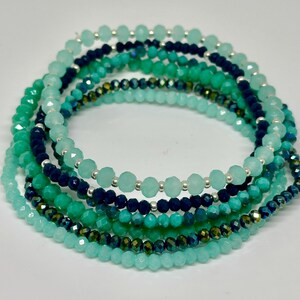 May include: A set of seven layered bracelets with turquoise, teal, and blue beads. The bracelets are made of glass and silver beads.