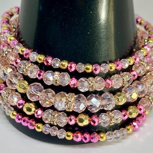 May include: A collection of beaded bracelets in pink, gold, and clear hues. The bracelets have faceted beads, gold-coloured accents, and decorative embellishments. They are displayed on a black cylindrical form.