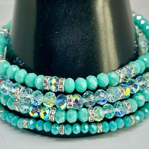 Könnte beinhalten: Ein Stapel von vier Armbändern mit türkisfarbenen und klaren Perlen. Die Armbänder haben facettierte Perlen und silberfarbene Abstandshalter mit klaren Kristallen. Die Armbänder werden auf einem schwarzen Ständer präsentiert.