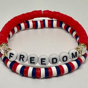 May include: Two stacked bracelets with red, white, and blue beads. The top bracelet is primarily red. The bottom bracelet has the word "FREEDOM" spelled out in white letter beads. Gold-colored accents are visible.