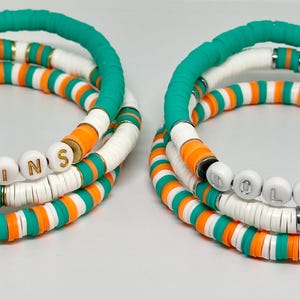 Könnte beinhalten: Zwei Sätze übereinanderliegender Armbänder mit weißen, orangefarbenen und türkisfarbenen Perlen. Die Perlen buchstabieren "DOLPHINS" in goldenen Buchstaben.