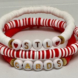 May include: Three bracelets with white and red beads. The top bracelet is white with gold letters spelling "STL" and a heart bead on each side. The middle bracelet is red and white striped. The bottom bracelet is red and white striped with gold letters spelling "CARDS".