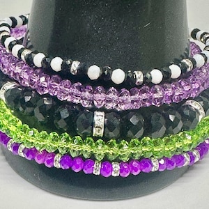 May include: Stack of beaded bracelets in various colors, including purple, black, green, and white. The bracelets feature faceted beads and silver-toned accents. The bracelets are stacked on a black display stand.