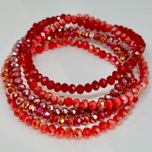 Könnte beinhalten: Ein Set aus fünf roten Kristall-Armbändern mit facettenreichem Finish. Die Armbänder sind übereinander gestapelt und erzeugen einen geschichteten Look.