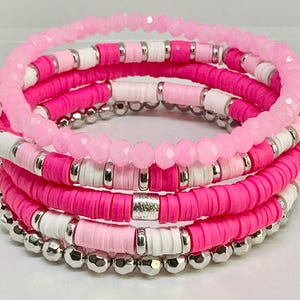Könnte beinhalten: Ein Set aus fünf rosafarbenen und weißen Perlenarmbändern mit silbernen Akzenten. Die Armbänder sind übereinander gestapelt und aus verschiedenen Materialien gefertigt, darunter Perlen, Kristalle und Metall.