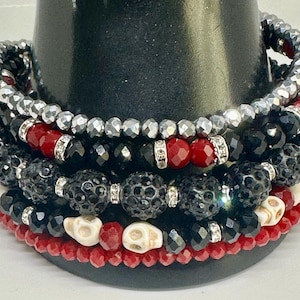 Può includere: Braccialetti impilati con una varietà di perline nere, rosse, argentate e bianche. I braccialetti includono perline sfaccettate, perline a teschio e dettagli in cristallo. I braccialetti sono esposti su un supporto nero.