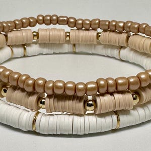 Könnte beinhalten: Ein Set aus drei Armbändern mit beigefarbenen und weißen Keramikperlen mit goldenen Akzenten. Die Armbänder sind übereinander gestapelt.