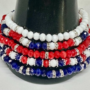 Può includere: Un set di sei braccialetti con perline rosse, bianche e blu. Le perline sono sfaccettate e hanno dettagli argentati. I braccialetti sono impilati su una superficie nera.