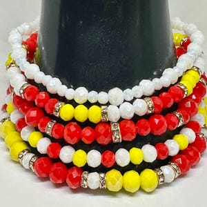 Könnte beinhalten: Ein Stapel Armbänder mit roten, weißen und gelben Perlen. Die Armbänder bestehen aus facettierten Perlen und sind mit goldfarbenen Abstandshaltern versehen. Die Armbänder werden auf einem schwarzen zylindrischen Objekt präsentiert.