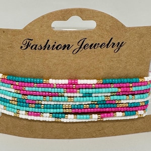 Pode incluir: Um conjunto de pulseiras de contas multicoloridas em rosa, turquesa, branco e dourado. As pulseiras estão empilhadas umas sobre as outras e são exibidas em um cartão marrom com o texto "Fashion Jewelry".