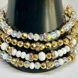 Pode incluir: Pilhas de pulseiras de contas em ouro, transparente e branco. As pulseiras apresentam uma mistura de contas facetadas e redondas, com detalhes de strass. As pulseiras são exibidas em um suporte cilíndrico preto.