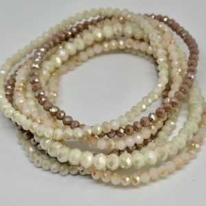 May include: A stack of beaded bracelets in various neutral colors, including cream, light pink, and brown. The bracelets are made of small, faceted beads, creating a sparkling effect. The bracelets are stacked together, showcasing the different colors and textures.