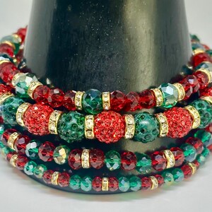 May include: A multi-strand stretch bracelet featuring alternating red and green faceted crystal beads, textured pavé-style spheres, and small gold-toned metal spacers. The beads are arranged in a repeating pattern around the circular band.
