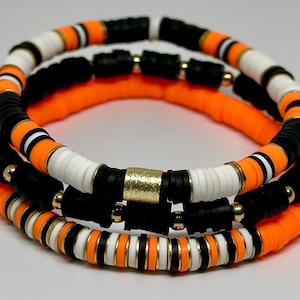 May include: Three layered bracelets with black, white, and orange beads. The beads are round and have a glossy finish. The bracelets are accented with gold beads.