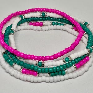 May include: A set of three beaded bracelets in white, pink, and teal colors. The bracelets are made of small, round beads and have silver accents.