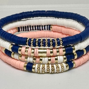 Puede incluir: Conjunto de cinco pulseras con cuentas de cerámica azul marino, blancas y rosas. Las pulseras están apiladas unas sobre otras.