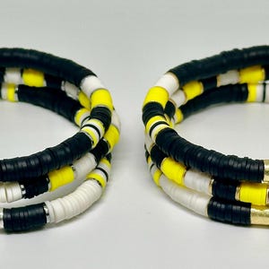 Puede incluir: Dos pulseras hechas de cuentas negras, blancas y amarillas. Las pulseras están apiladas una encima de la otra.