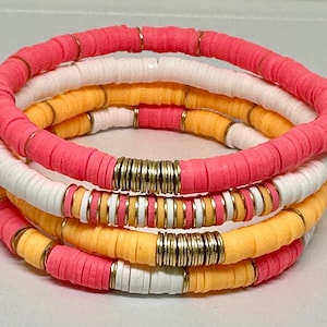 May include: A set of four colourful bracelets made of stacked, round, flat beads. The bracelets are pink, white, and orange with gold accents.