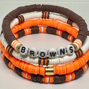 May include: Stack of four beaded bracelets in brown, white, and orange. The bracelets have the word "BROWNS" spelled out in white letter beads. The bracelets are made of small, flat, round beads.