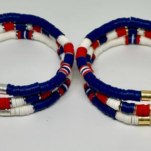 May include: Two sets of layered bracelets with blue, red, and white beads. The bracelets are made of small, round beads and have a gold accent.