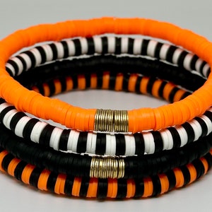 May include: Stack of four bracelets in orange, black, and white. The bracelets are made of small, flat, round beads. Two bracelets are orange, one is black, and one has black and white stripes. The bracelets are held together with gold-colored metal.