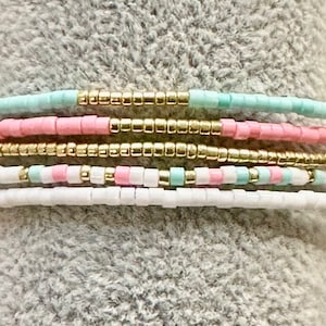 May include: A set of six beaded bracelets in pink, light blue, and gold. The bracelets are stacked on top of each other.