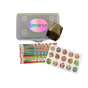 May include: A clear plastic pencil box with the name "Emmerson" on a holographic sticker. Includes a black pencil sharpener, a stack of pencils with "Emmerson" printed on them, and a sheet of Mario-themed stickers.